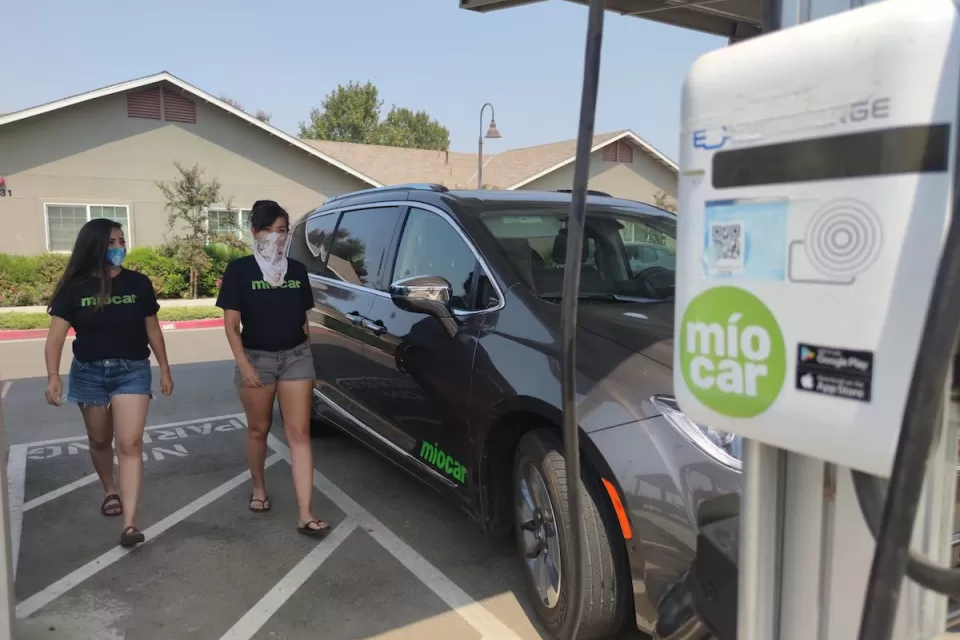 Black SUV charging at Miocar electric car sharing station with two women walking towards it.